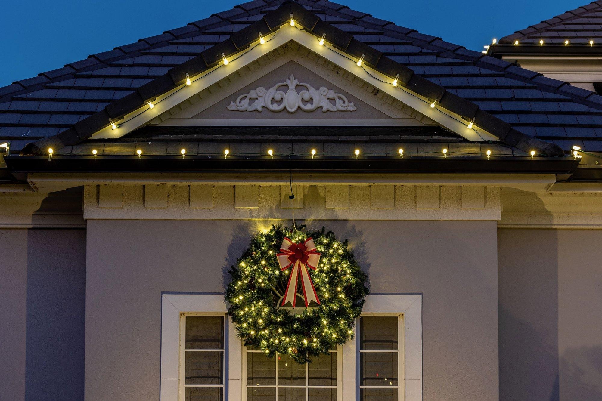 Glowing holiday wreath with a red bow above a window, framed by warm roofline lights