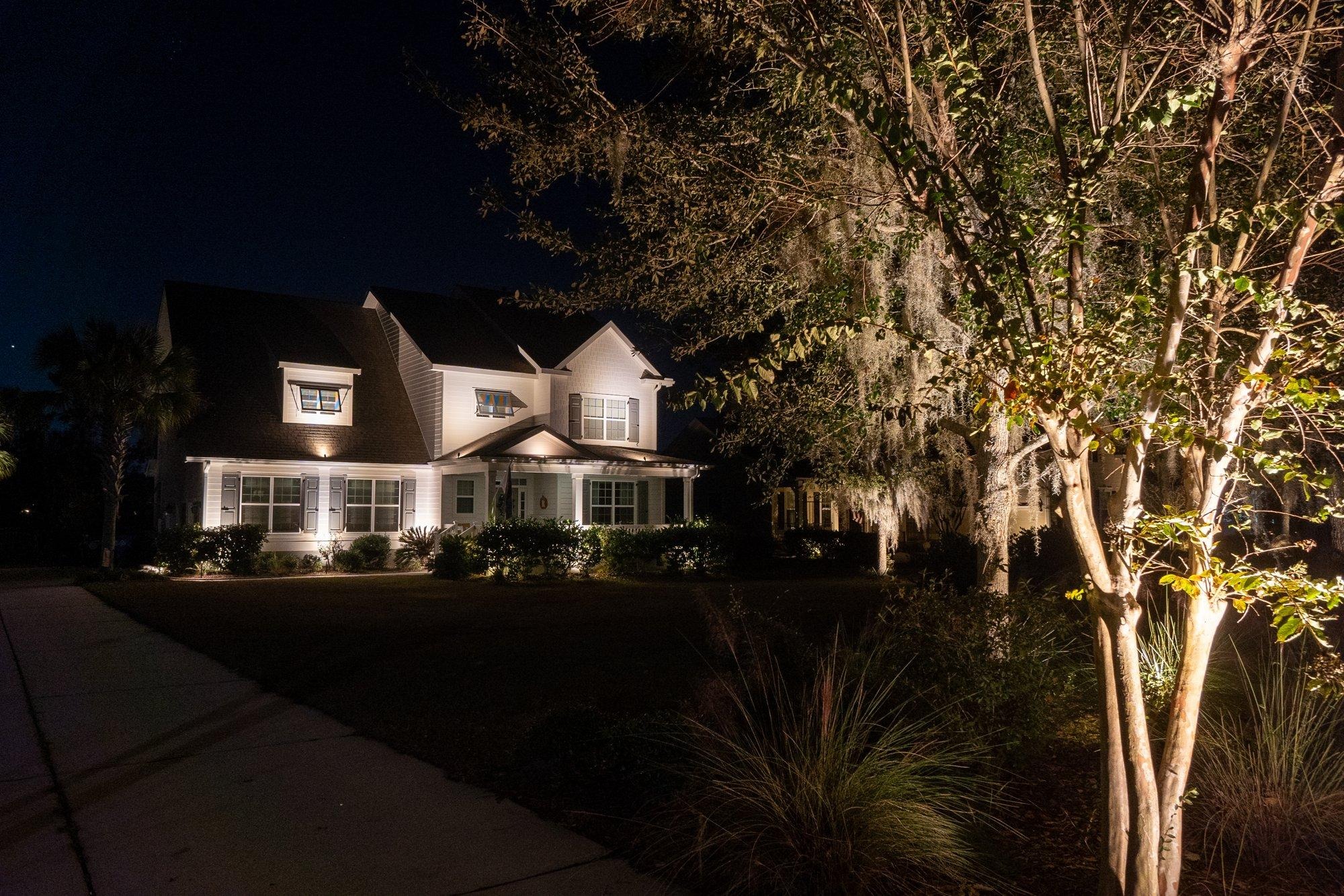 Classic white two-story home glowing with warm architectural lighting beside a softly uplit oak