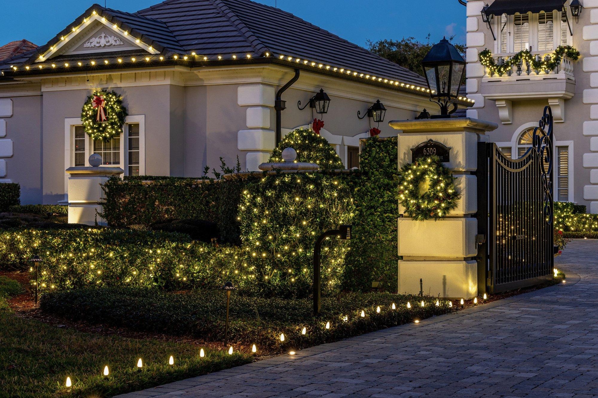 Lit garland on a gated entry with glowing hedges, wreaths, and holiday lights lining the driveway