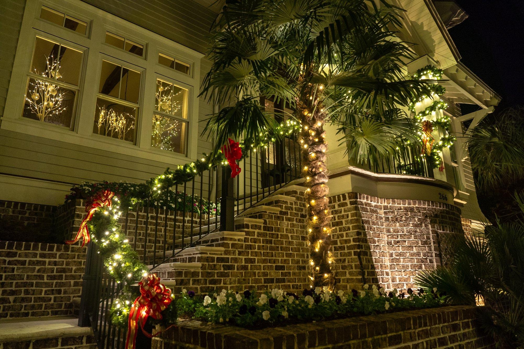 Brick front steps wrapped in lit garland with red bows and wreaths, framed by palms and warm porch lighting