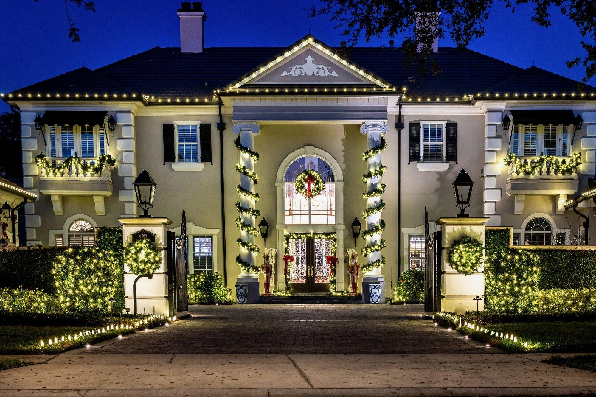 Grand home dressed for the holidays with wreaths in every window, lit garland, glowing hedges, and warm roofline lighting