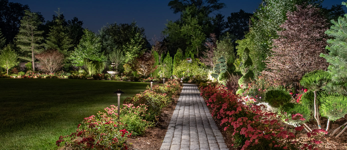 A brick path with flowers and trees at nightDescription automatically generated